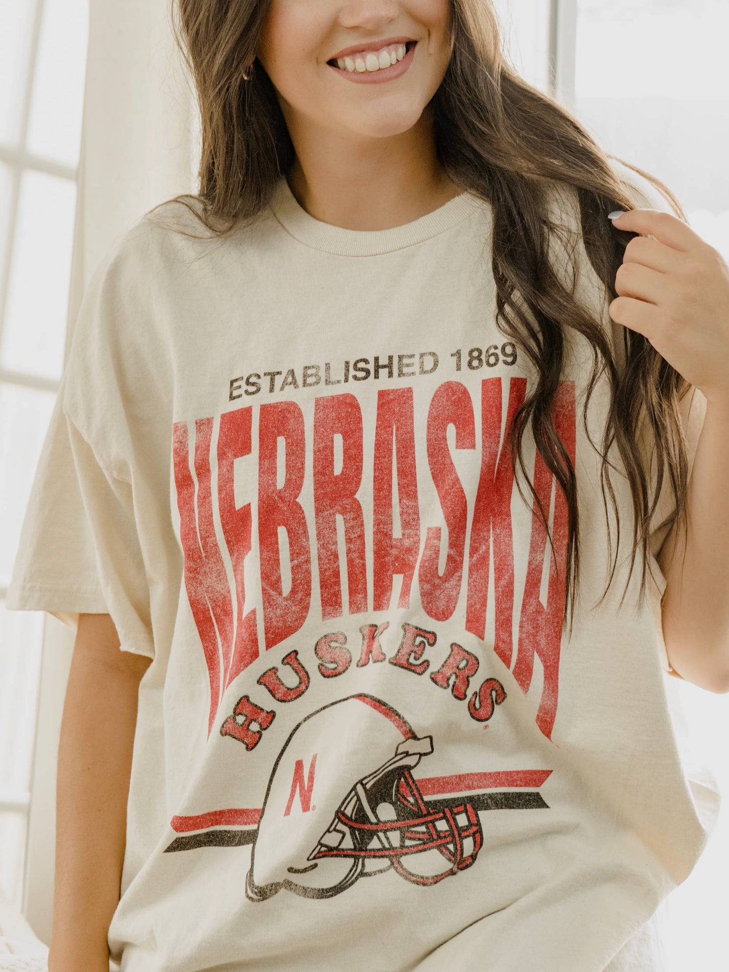 styled view of Nebraska Established Date Helmet Off White Thrifted Tee, 100% preshrunk cotton, unisex fit, meaningfully distressed, and officially licensed with unique colors and distressing.