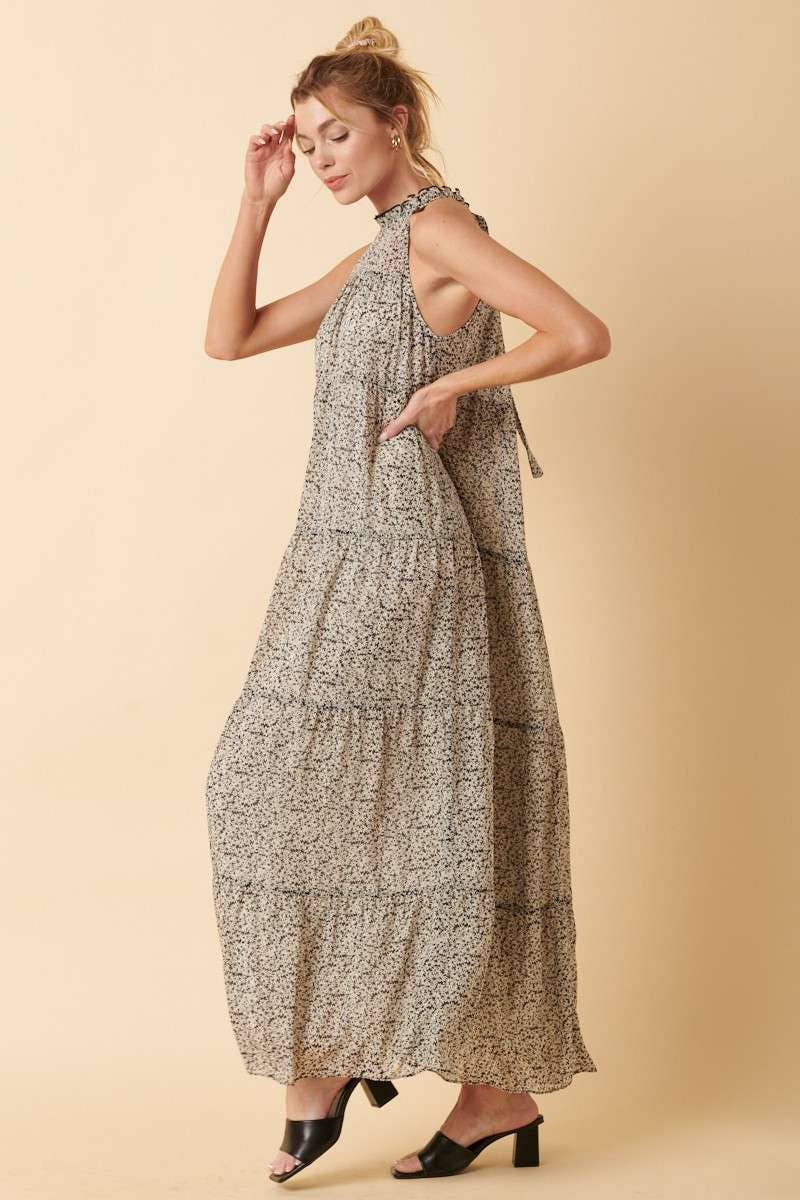 side view of Sally Flower Halter Maxi Dress with flower print on chiffon fabric, ruffle mock neck, tie back, sleeveless halter design, and tiered detail. Maxi length for an elegant silhouette.