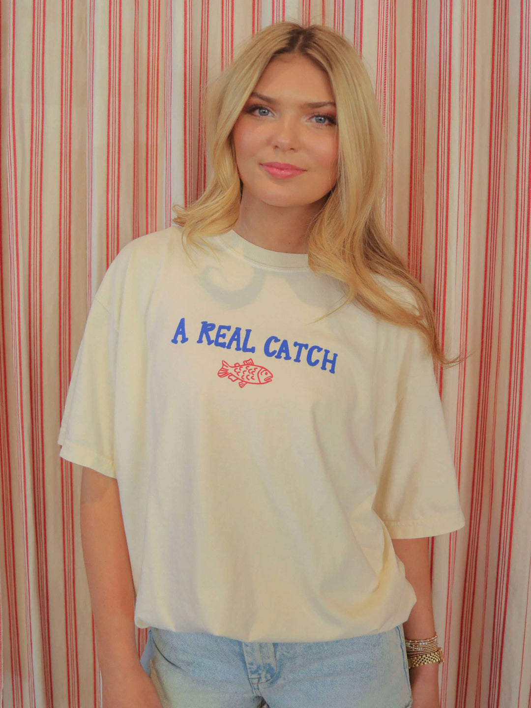 model showing front view of Americana-inspired graphic tee with 'Catch of the Day' text. Features a fun, lake-themed graphic perfect for boaters and fishing enthusiasts.
