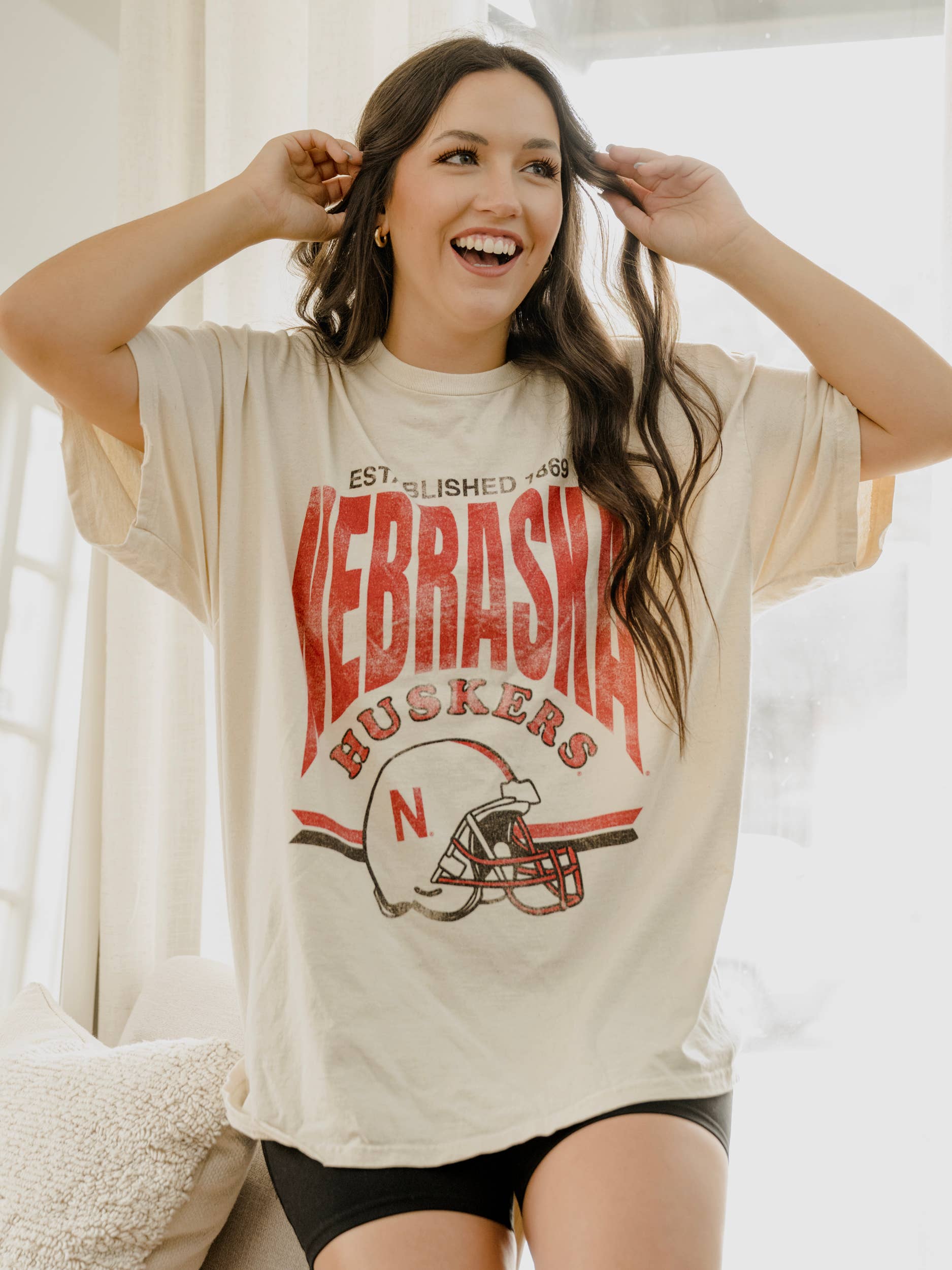 arms up view of Nebraska Established Date Helmet Off White Thrifted Tee, 100% preshrunk cotton, unisex fit, meaningfully distressed, and officially licensed with unique colors and distressing.