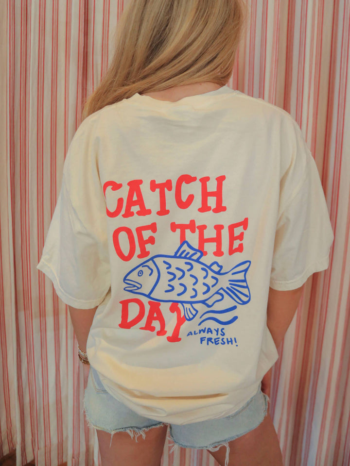 model showing back view of Americana-inspired graphic tee with 'Catch of the Day' text. Features a fun, lake-themed graphic perfect for boaters and fishing enthusiasts.