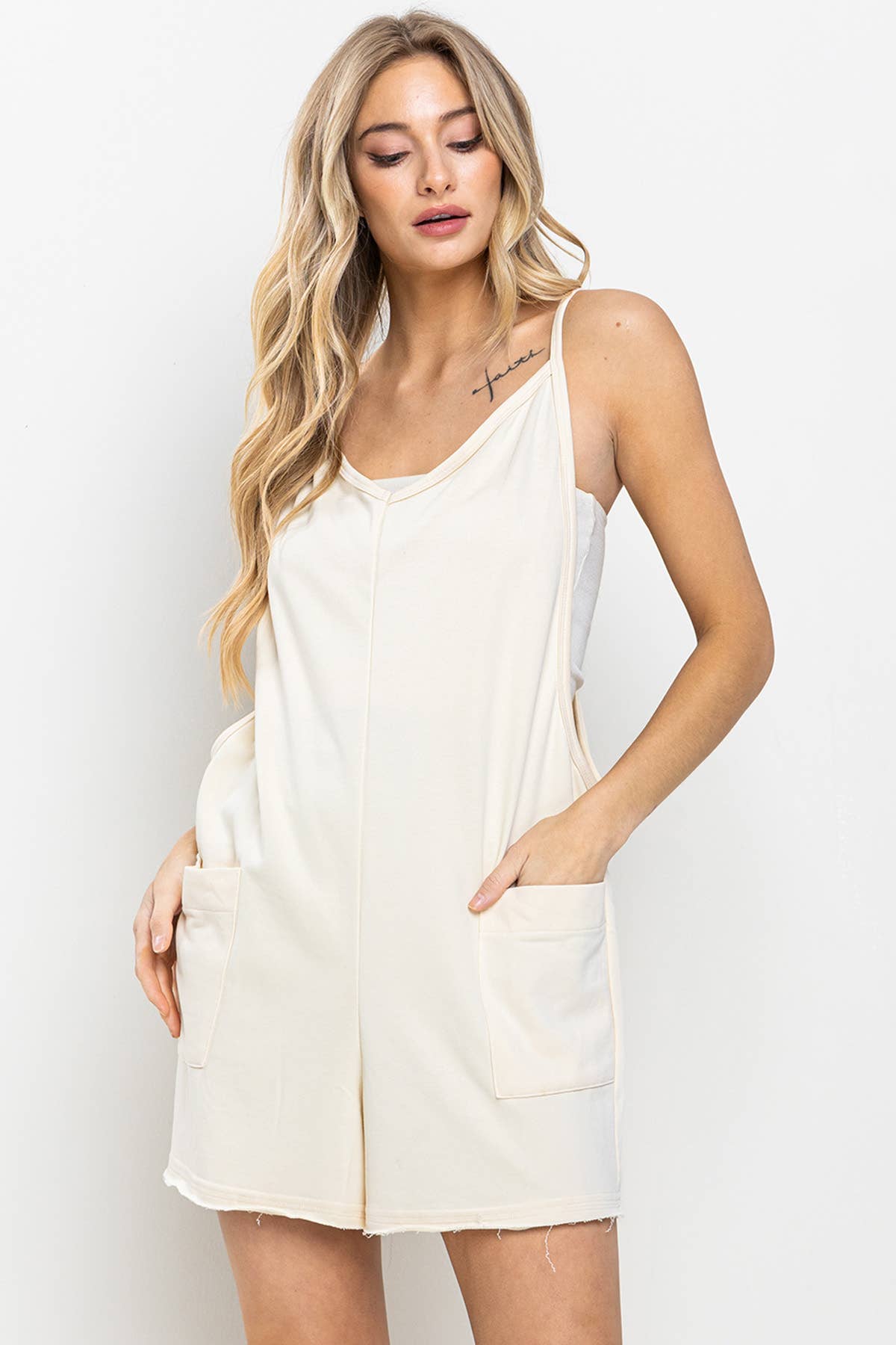 pocket view of Cream V-Neck Romper with raw edge details, spaghetti straps, deep armholes, front flap pockets, and a back zipper. Made from solid stretch fabric with a garment-washed finish.