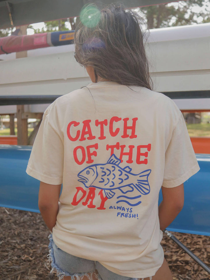 back view of Americana-inspired graphic tee with 'Catch of the Day' text. Features a fun, lake-themed graphic perfect for boaters and fishing enthusiasts.