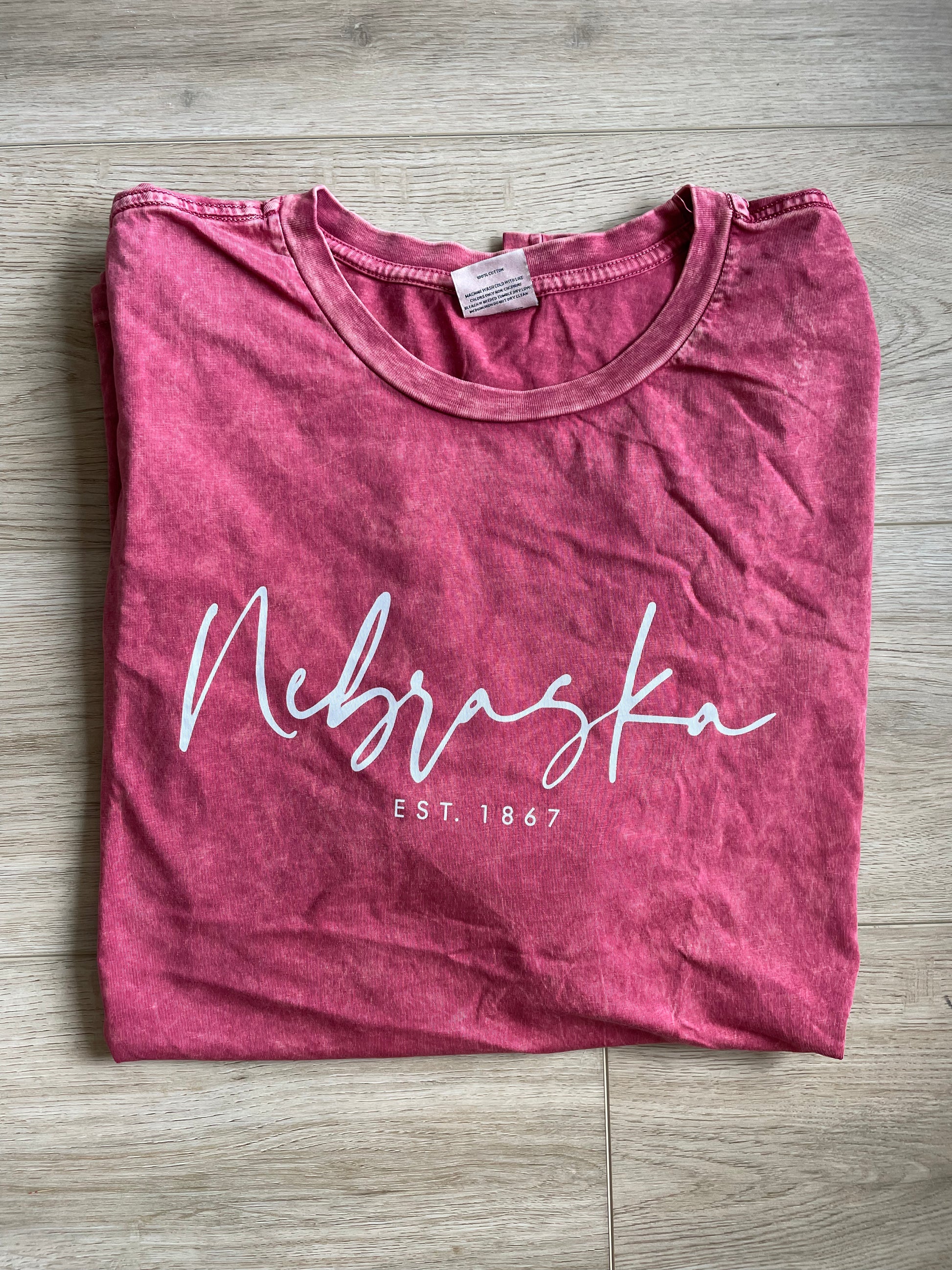 folded view of NEBRASKA EST. 1867 Graphic Plus Size Mineral Washed Top in cardinal red. Made from 100% premium cotton jersey, featuring a relaxed fit and screen printed design with water-based ink. Each top is uniquely dyed, offering a distinctive look.