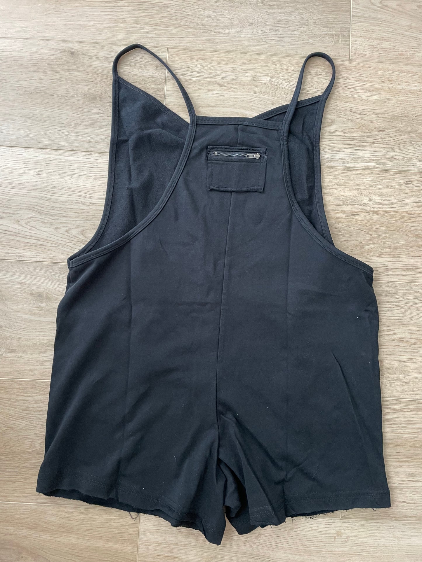 back view of Black V-neck romper featuring raw edge details, solid stretch fabric, and a garment-washed finish. Designed with a deep armhole, spaghetti straps, front flap pockets, and a back zipper detail.