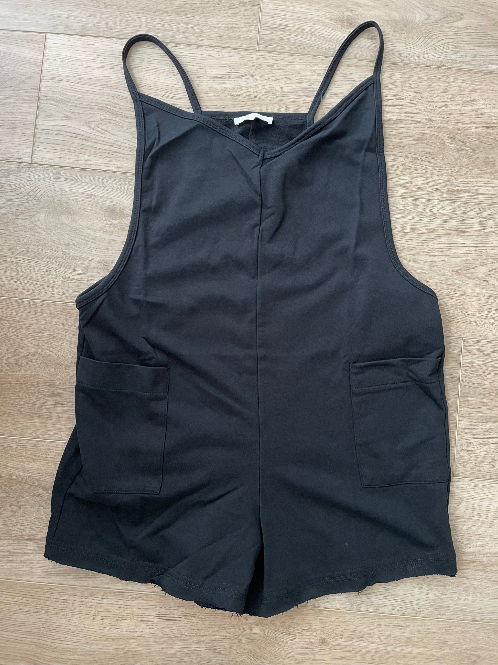 real view of Black V-neck romper featuring raw edge details, solid stretch fabric, and a garment-washed finish. Designed with a deep armhole, spaghetti straps, front flap pockets, and a back zipper detail.