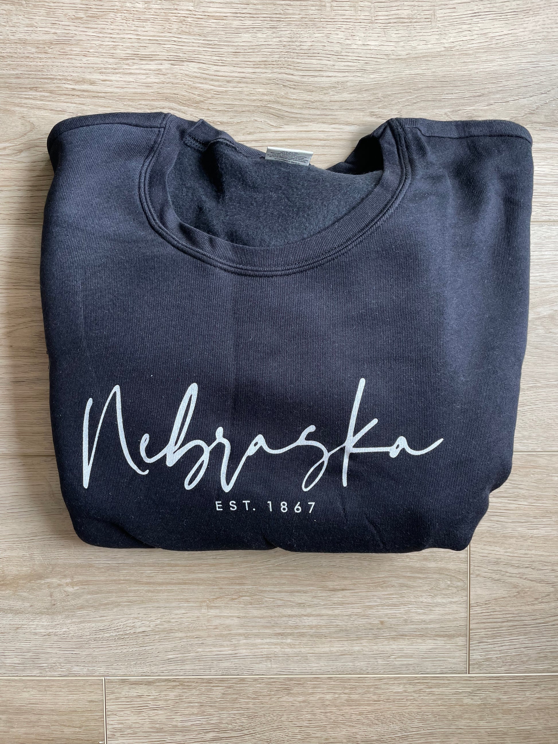 folded view of "NEBRASKA EST. 1867" plus graphic sweatshirt in unisex crewneck style. Made from soft fleece with ribbed cuffs and waistband, ideal for daily wear and layering. Refer to size chart for measurements.