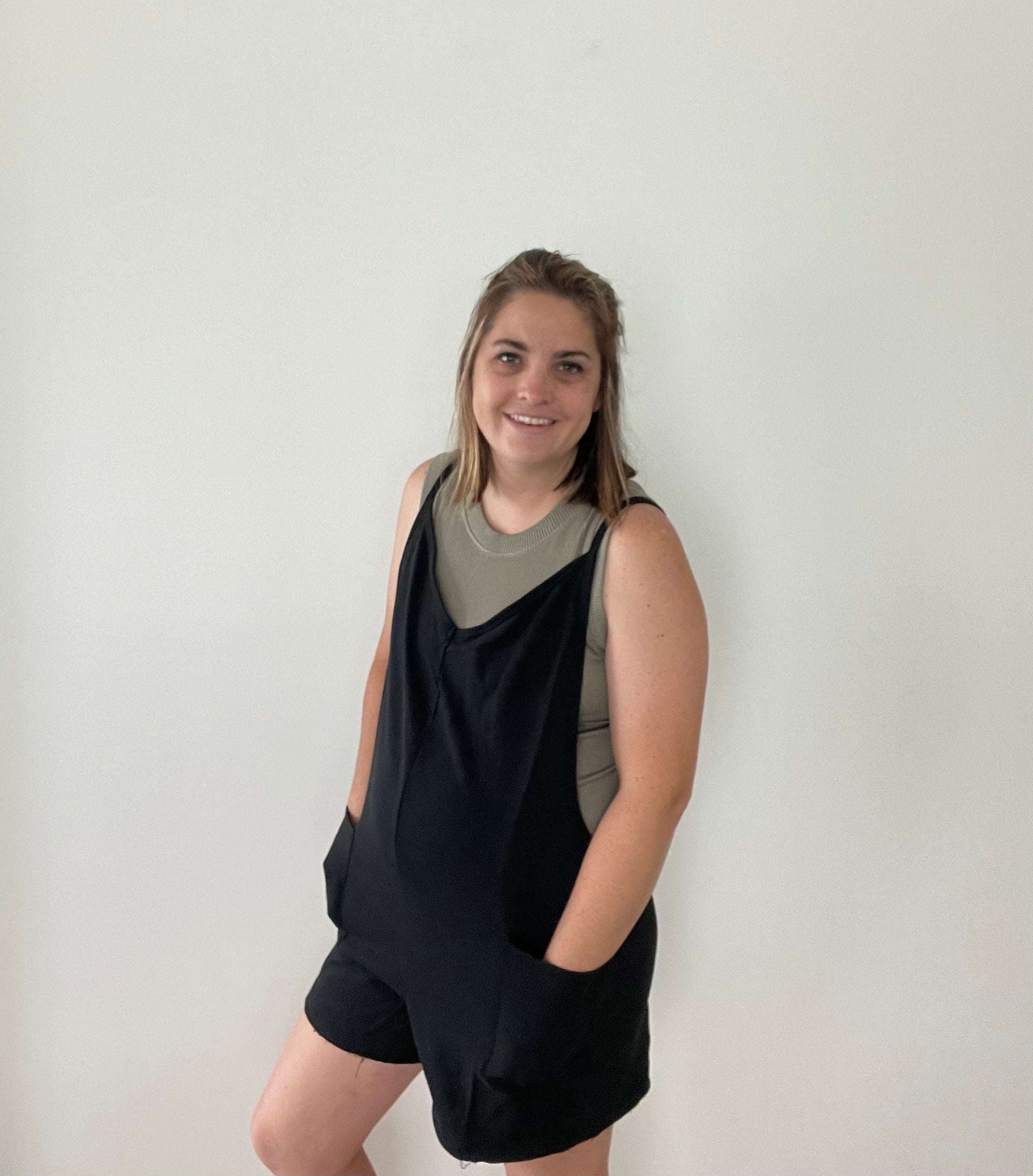 model view of Black V-neck romper featuring raw edge details, solid stretch fabric, and a garment-washed finish. Designed with a deep armhole, spaghetti straps, front flap pockets, and a back zipper detail.