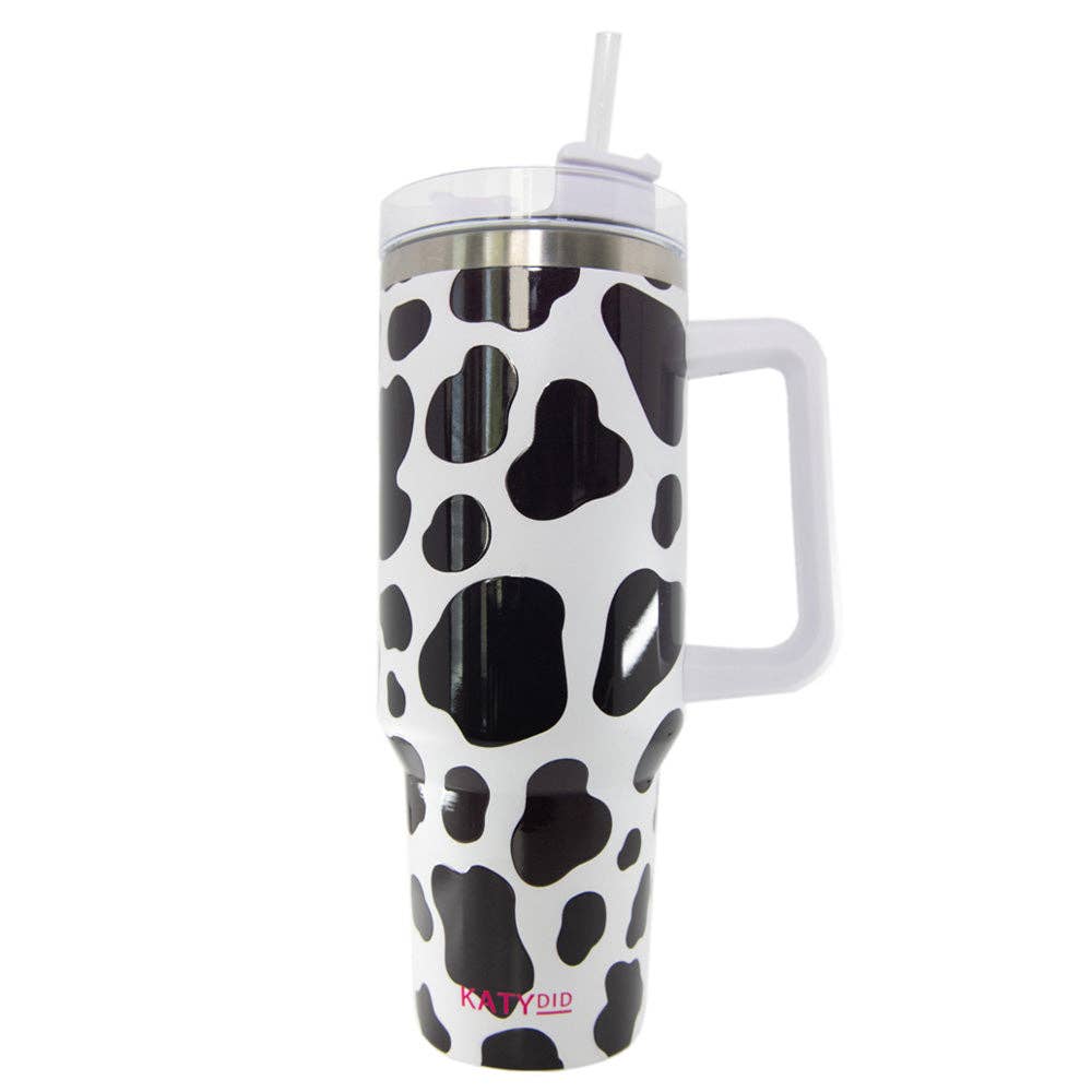 Katydid Wholesale Cow Print 40 oz Tumbler featuring a black and white cow print pattern, stainless steel interior, and a reusable plastic straw. Includes an adjustable straw cover and a screw-on plastic top with a straw opening. Designed with an easy-to-hold handle and fits in car cup holders.